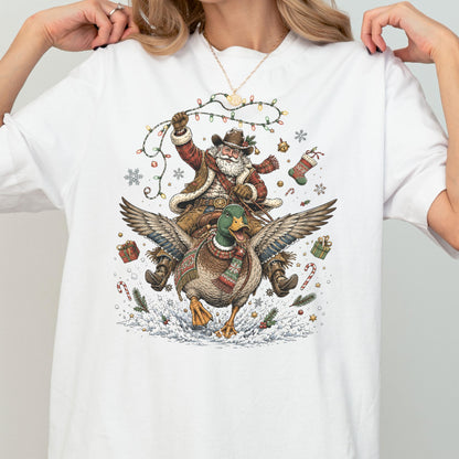 May include: A white t-shirt featuring a graphic illustration of Santa Claus wearing a cowboy hat and vest, riding a mallard duck. The scene includes Christmas lights, a stocking, gift boxes, candy canes, and snowflakes on a snowy ground.