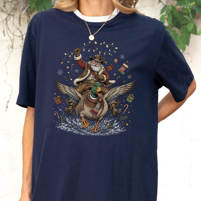 May include: A navy blue short-sleeve t-shirt featuring a graphic print of a cowboy Santa Claus riding a flying mallard duck. The design includes Christmas lights, snowflakes, candy canes, a stocking, and gift boxes surrounding the central figures.