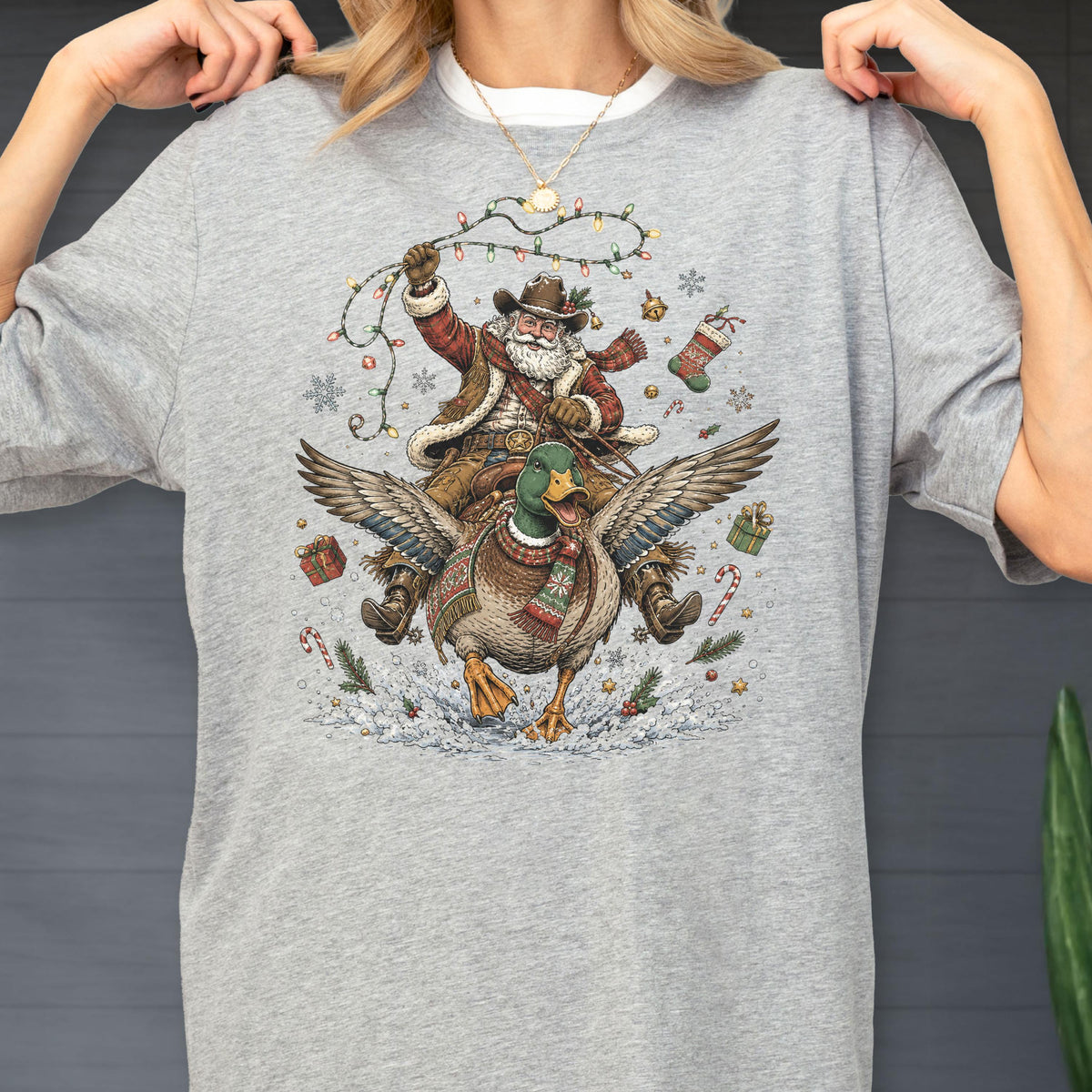 May include: A heather gray t-shirt featuring a graphic illustration of Santa Claus wearing a cowboy hat and scarf, riding a mallard duck. The design includes Christmas lights, snowflakes, gift boxes, and candy canes surrounding the central figures.