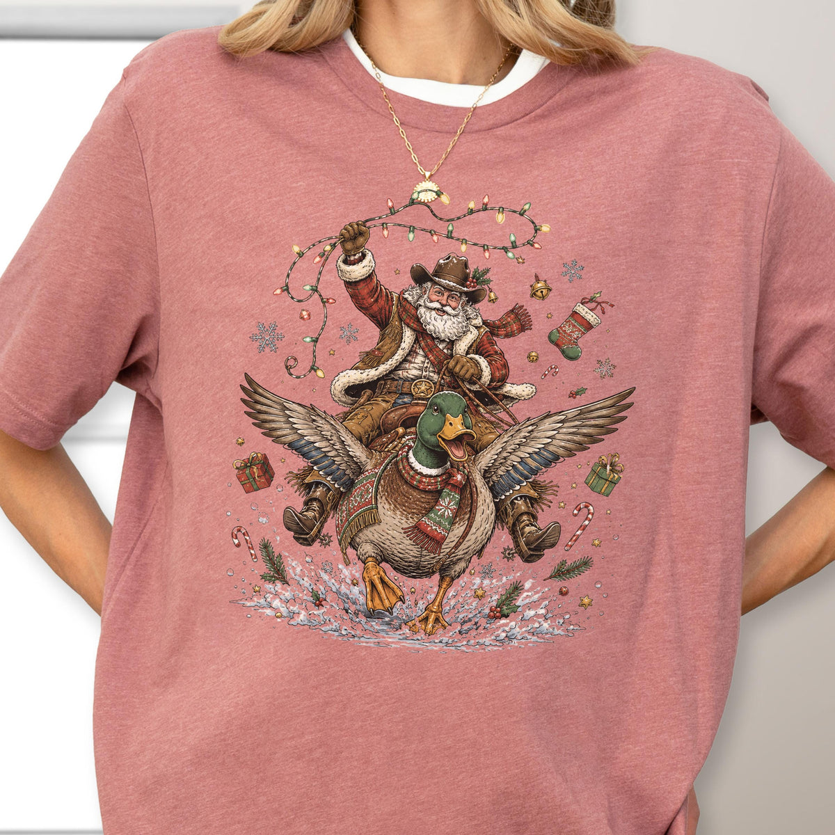 May include: A dusty rose t-shirt featuring a graphic of Santa Claus wearing a cowboy hat and riding a mallard duck. Santa holds a string of Christmas lights, surrounded by floating snowflakes, gift boxes, a stocking, and candy canes.