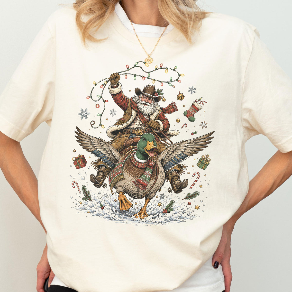 May include: A cream-colored t-shirt featuring a graphic illustration of Santa Claus wearing a cowboy hat and riding a flying mallard duck. Santa holds a string of colorful Christmas lights, surrounded by snowflakes, gifts, and candy canes.
