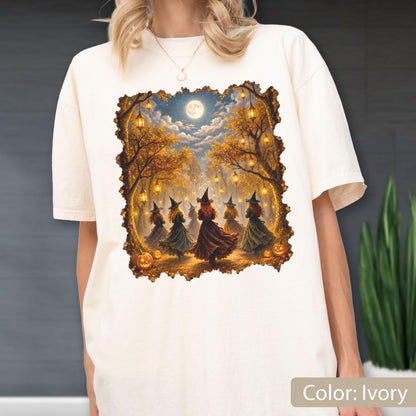 May include: An ivory short-sleeve t-shirt featuring a rectangular graphic print of five figures wearing pointed hats and long dresses dancing in a forest at night. The scene includes glowing lanterns hanging from trees, a full moon, and pumpkins.