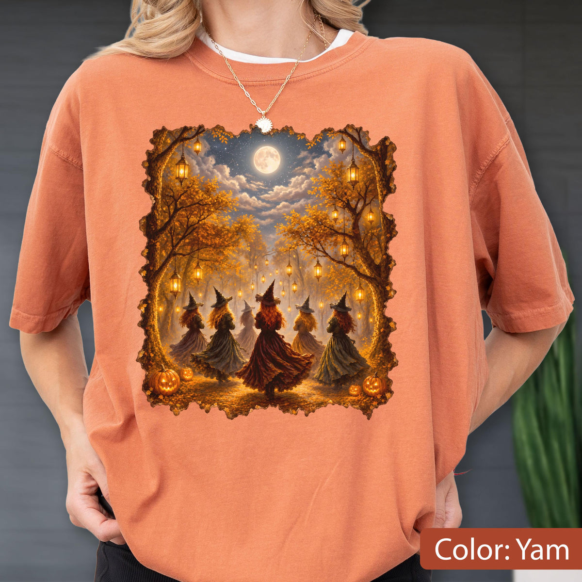 May include: A yam-colored t-shirt featuring a rectangular graphic print of several figures in pointed hats and long dresses dancing in a forest at night. The scene includes glowing lanterns hanging from autumn trees, a full moon, and orange pumpkins.