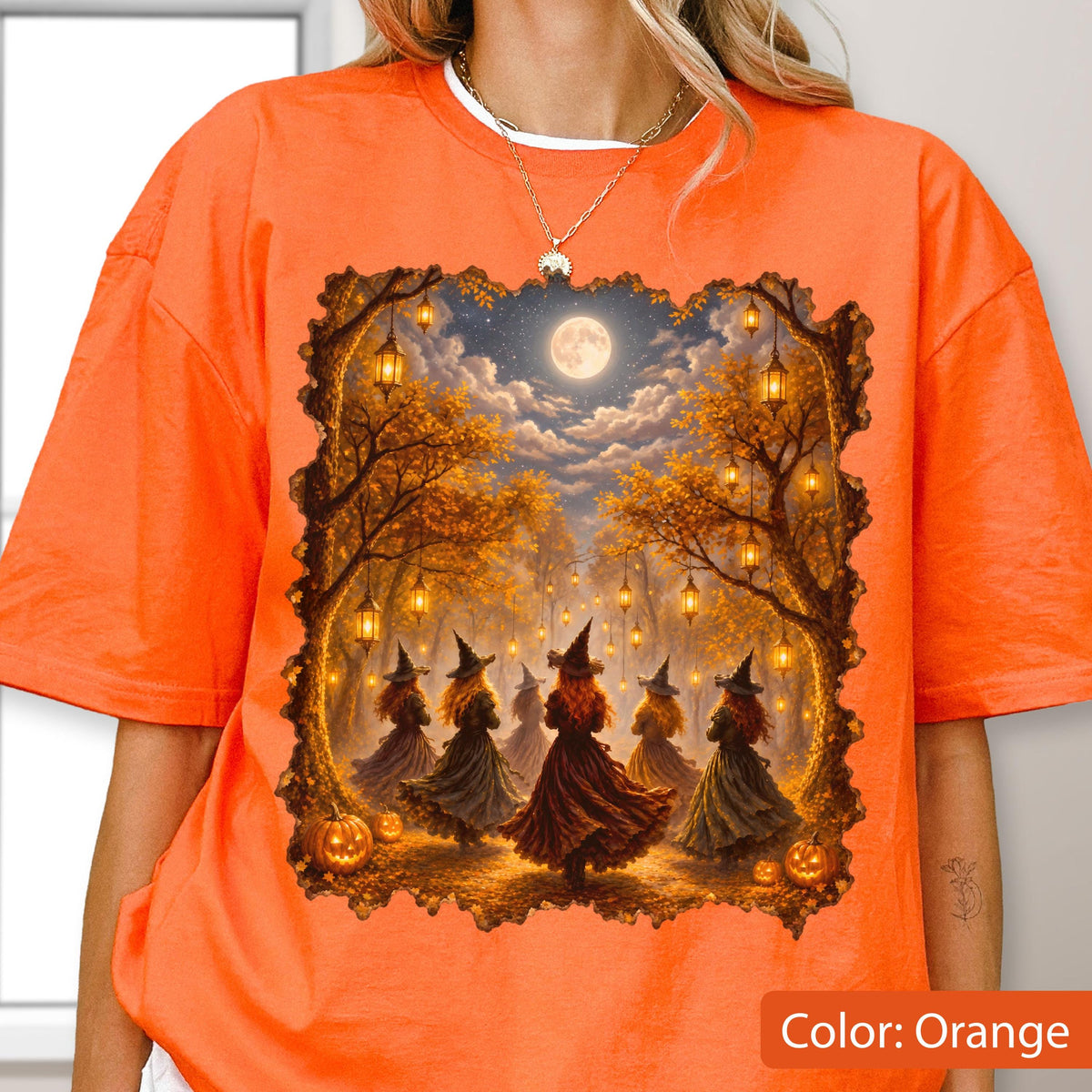 May include: A bright orange t-shirt featuring a rectangular graphic print of five figures wearing pointed hats and long dresses standing in a forest clearing under a full moon, surrounded by hanging lanterns and pumpkins on the ground.