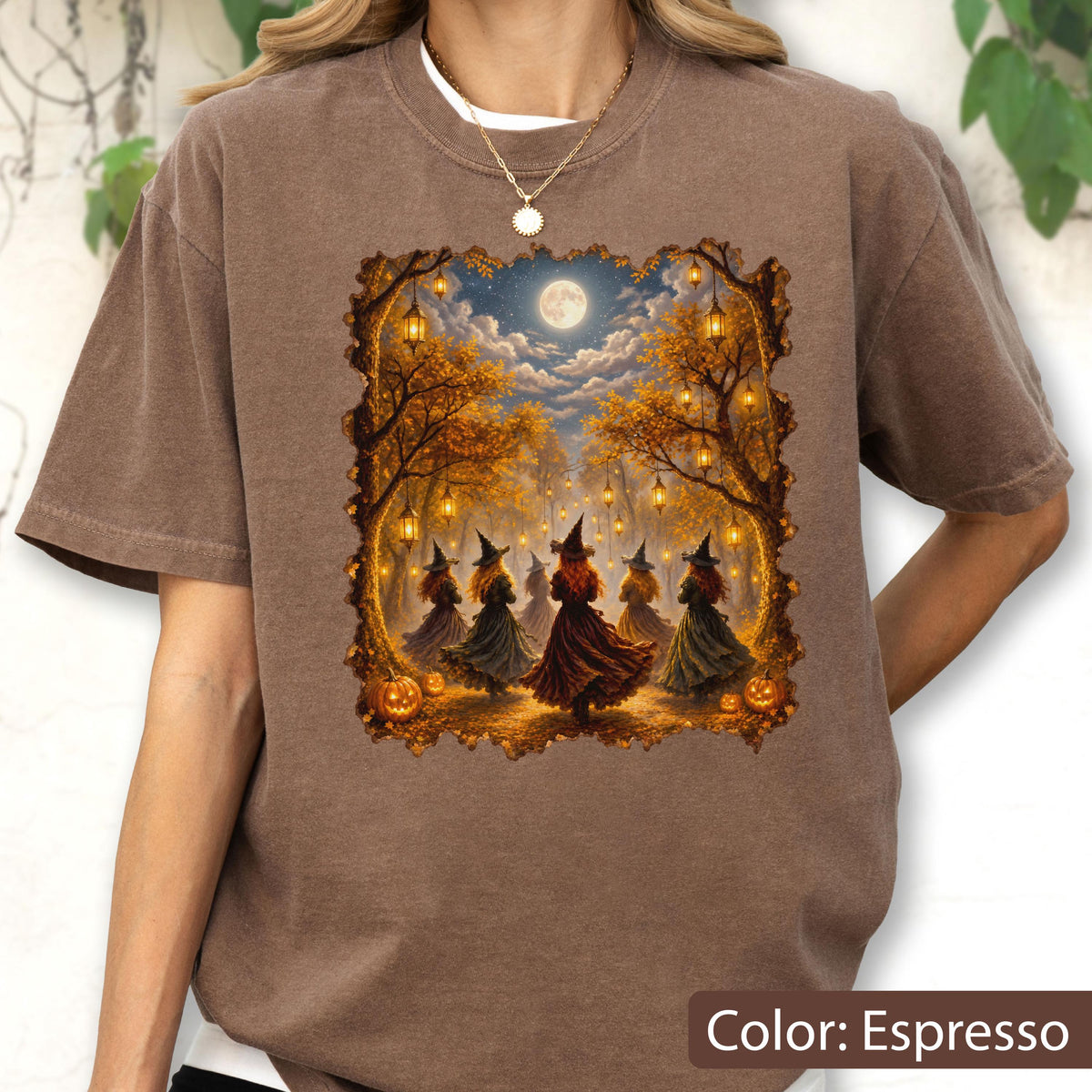 May include: An espresso-colored short-sleeve t-shirt featuring a rectangular graphic print of five figures wearing pointed hats and long dresses dancing in a forest at night under a full moon, surrounded by hanging lanterns and orange pumpkins.