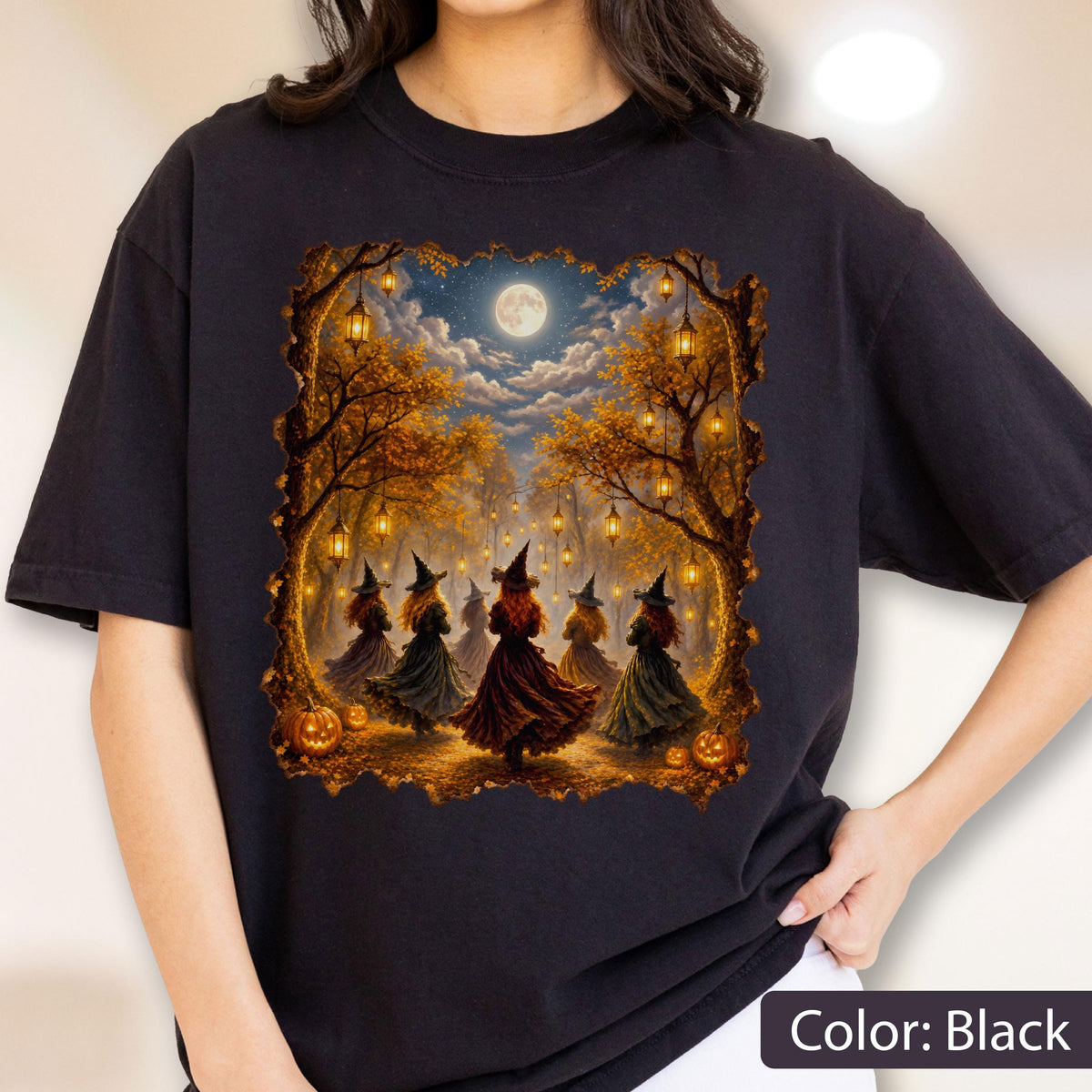 May include: A black t-shirt featuring a graphic print of several figures in pointed hats and long dresses dancing in a forest at night. The scene includes glowing lanterns hanging from autumn trees, a full moon in the sky, and orange pumpkins.