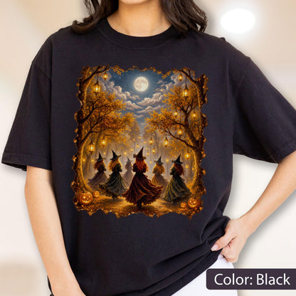 May include: A black t-shirt featuring a graphic print of several figures in pointed hats and long dresses dancing in a forest at night. The scene includes glowing lanterns hanging from autumn trees, a full moon in the sky, and orange pumpkins.