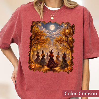 May include: A crimson short-sleeve t-shirt featuring a graphic print of several figures in witch hats and long dresses dancing in a forest at night. The scene includes hanging lanterns, autumn trees, a full moon, and pumpkins on the ground.