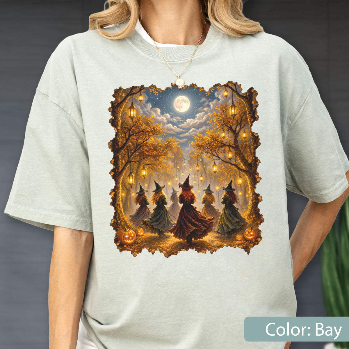 May include: A light sage green t-shirt featuring a rectangular graphic print of several figures in pointed hats and long dresses dancing in a forest at night. The scene includes glowing lanterns hanging from trees, a full moon, and orange pumpkins.