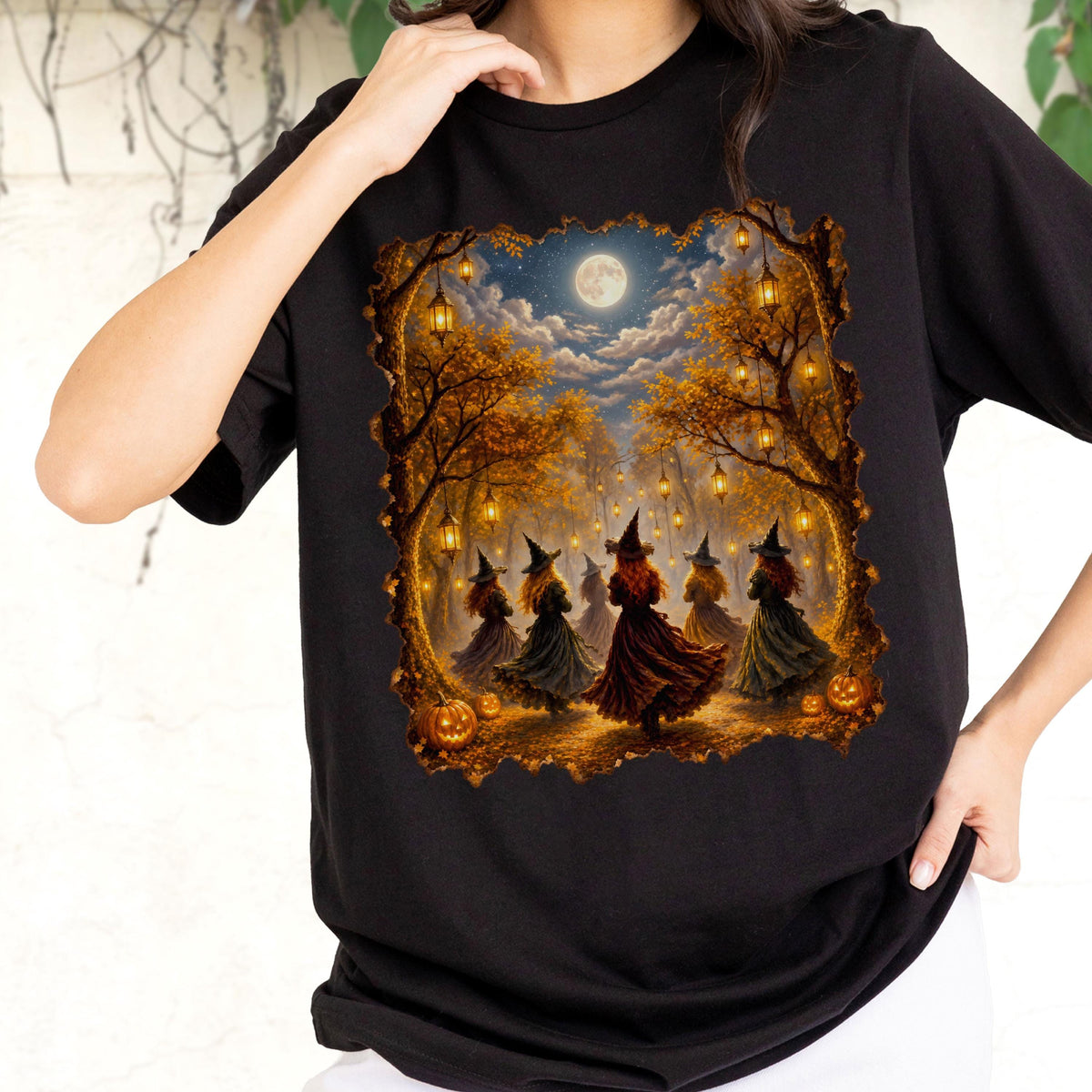 May include: A black t-shirt featuring a rectangular graphic print of several figures wearing pointed hats and long dresses dancing in a forest at night. The scene includes glowing lanterns hanging from autumn trees, a full moon, and orange pumpkins.