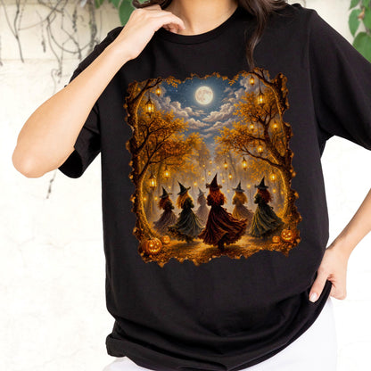 May include: A black t-shirt featuring a rectangular graphic print of several figures wearing pointed hats and long dresses dancing in a forest at night. The scene includes glowing lanterns hanging from autumn trees, a full moon, and orange pumpkins.