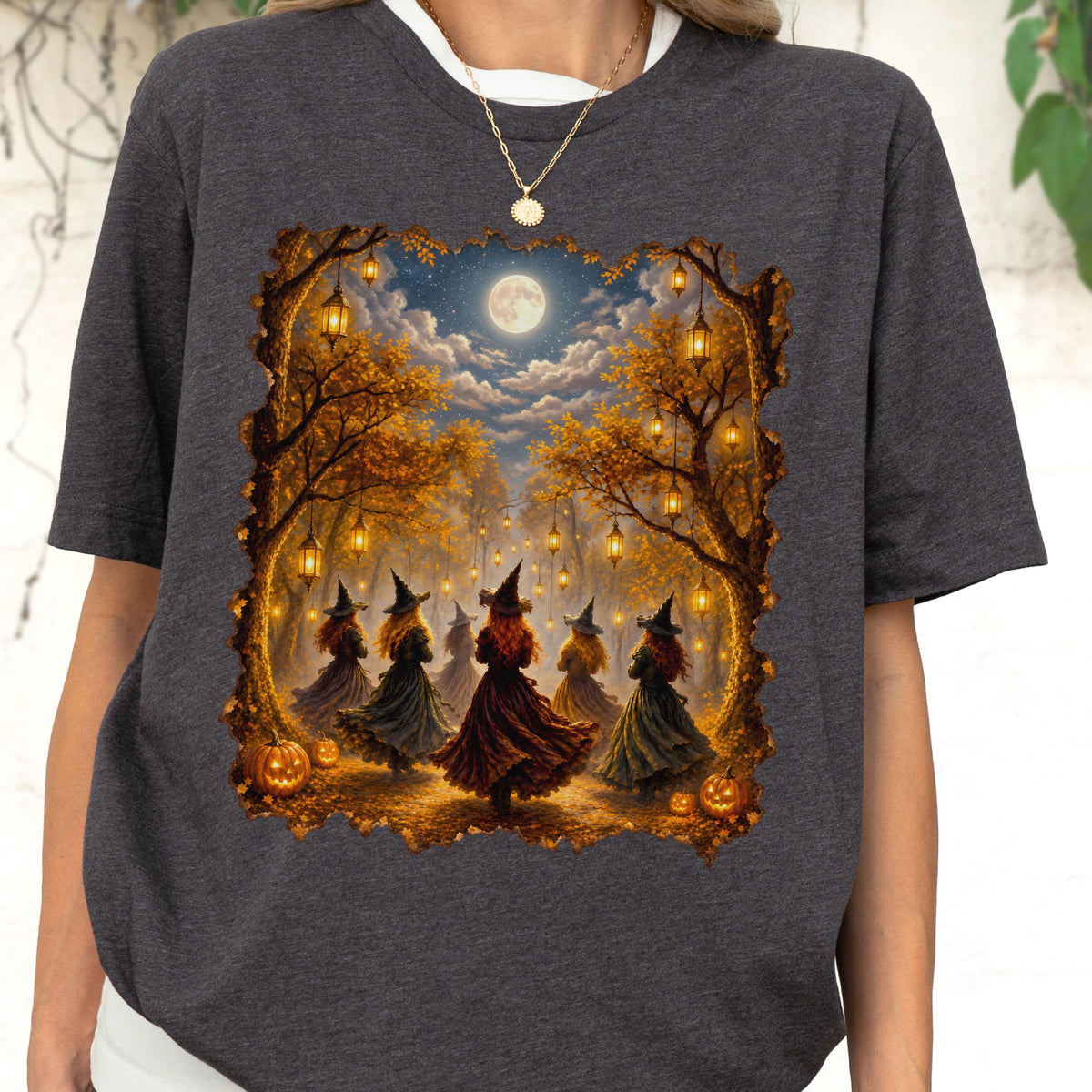 May include: A dark heather gray t-shirt featuring a graphic print of several figures in pointed hats and long dresses dancing in a forest at night. The scene includes glowing lanterns hanging from trees, a full moon, and orange pumpkins on the ground.