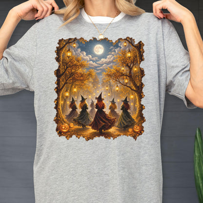 May include: A heather gray t-shirt featuring a rectangular graphic print of five figures wearing pointed hats and long dresses standing in an autumn forest at night. The scene includes hanging lanterns, glowing pumpkins, and a full moon in the sky.
