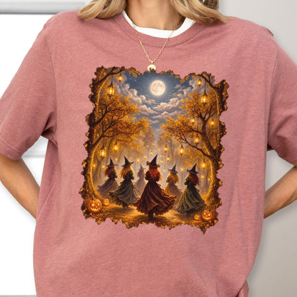 May include: A dusty rose short-sleeved t-shirt featuring a rectangular graphic print of six figures in pointed hats dancing in an autumn forest at night. The scene includes glowing lanterns, orange pumpkins, and a full moon under a cloudy sky.