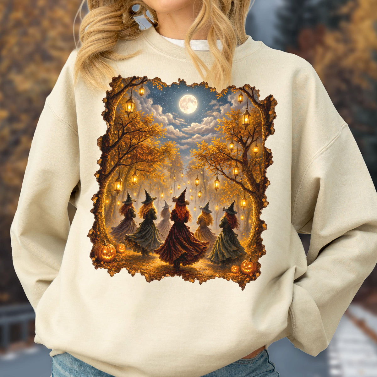 May include: A beige crewneck sweatshirt featuring a rectangular graphic print of several figures in pointed hats and long dresses dancing in a forest at night. The scene includes glowing lanterns hanging from trees, a full moon, and orange pumpkins.