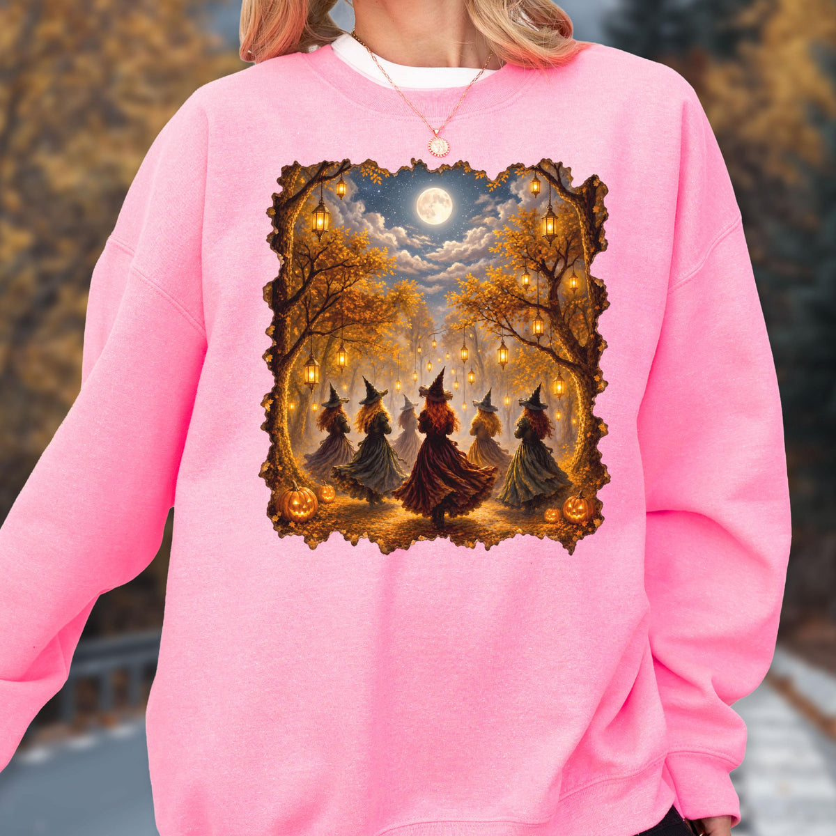 May include: A bright pink long-sleeve sweatshirt featuring a rectangular graphic print of five figures in pointed hats and dresses dancing in a forest at night. The scene includes glowing hanging lanterns, autumn trees, a full moon, and pumpkins.