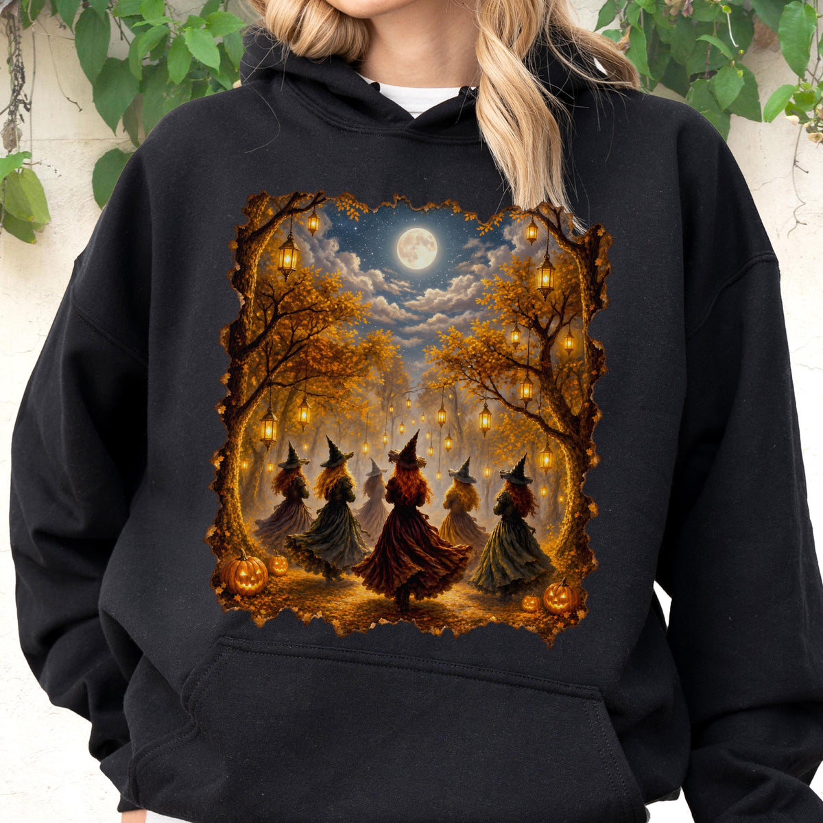 May include: A black hooded sweatshirt featuring a rectangular graphic print of several figures wearing pointed hats and long dresses dancing in a forest at night. The scene includes glowing lanterns hanging from autumn trees, pumpkins, and a full moon.