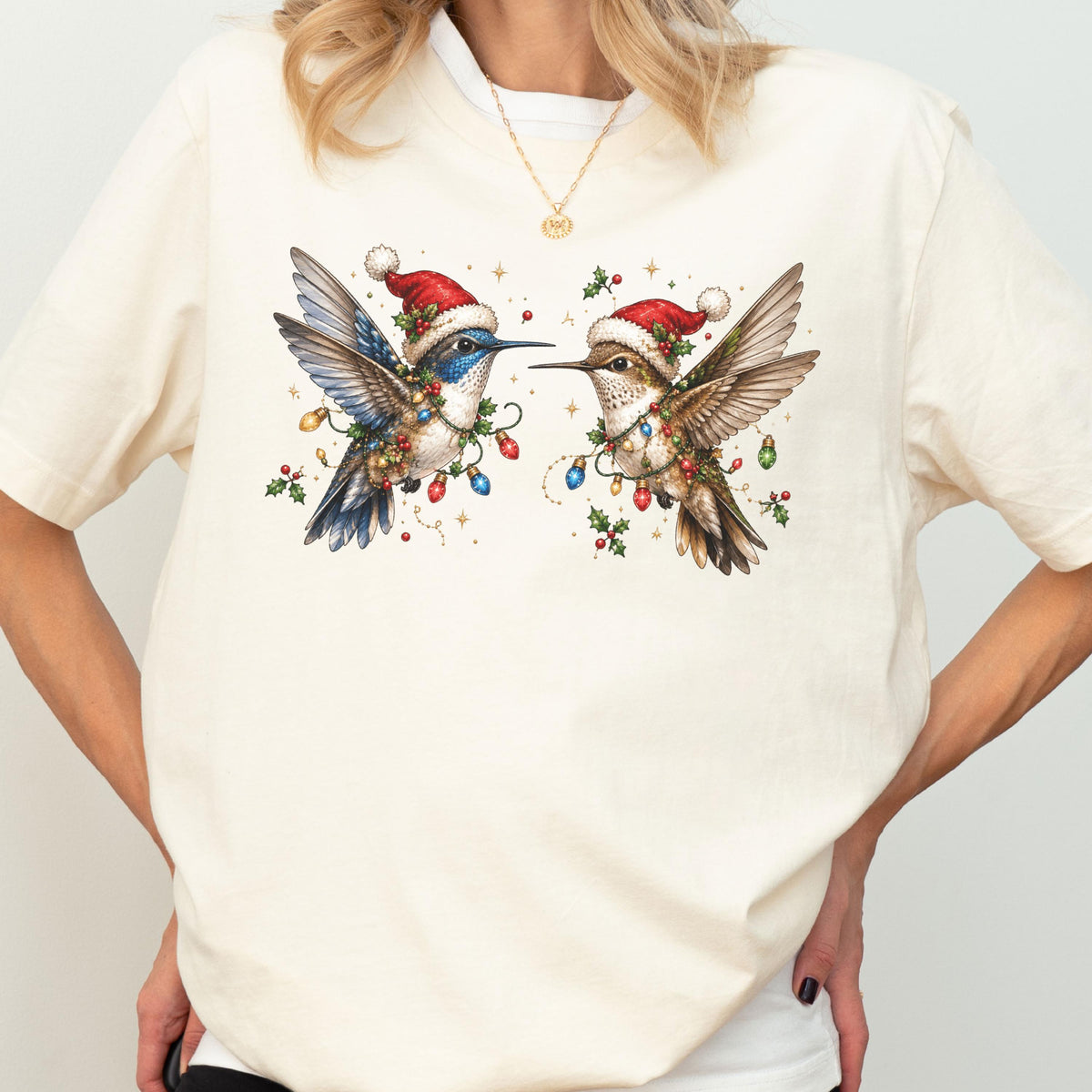 May include: A cream-colored t-shirt featuring a graphic illustration of two hummingbirds facing each other. Both birds wear red Santa hats and are adorned with colorful Christmas light strings and green holly leaves with red berries.