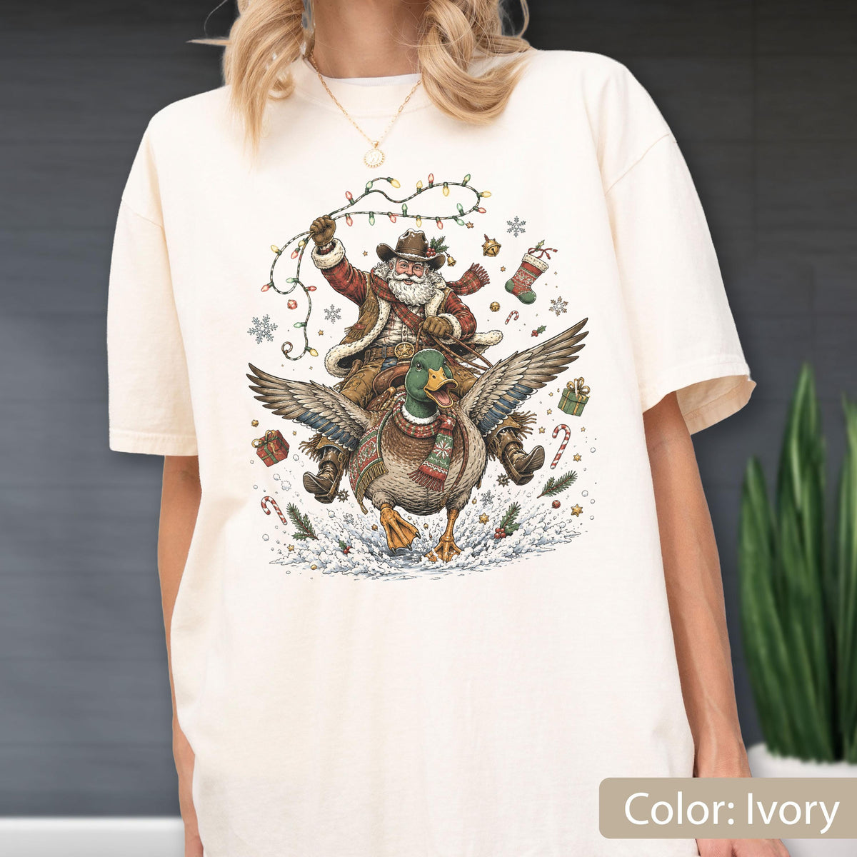 May include: An ivory t-shirt featuring a graphic of Santa Claus wearing a cowboy hat and riding a flying mallard duck. Santa holds a string of colorful Christmas lights. The design includes snowflakes, wrapped gifts, and candy canes.