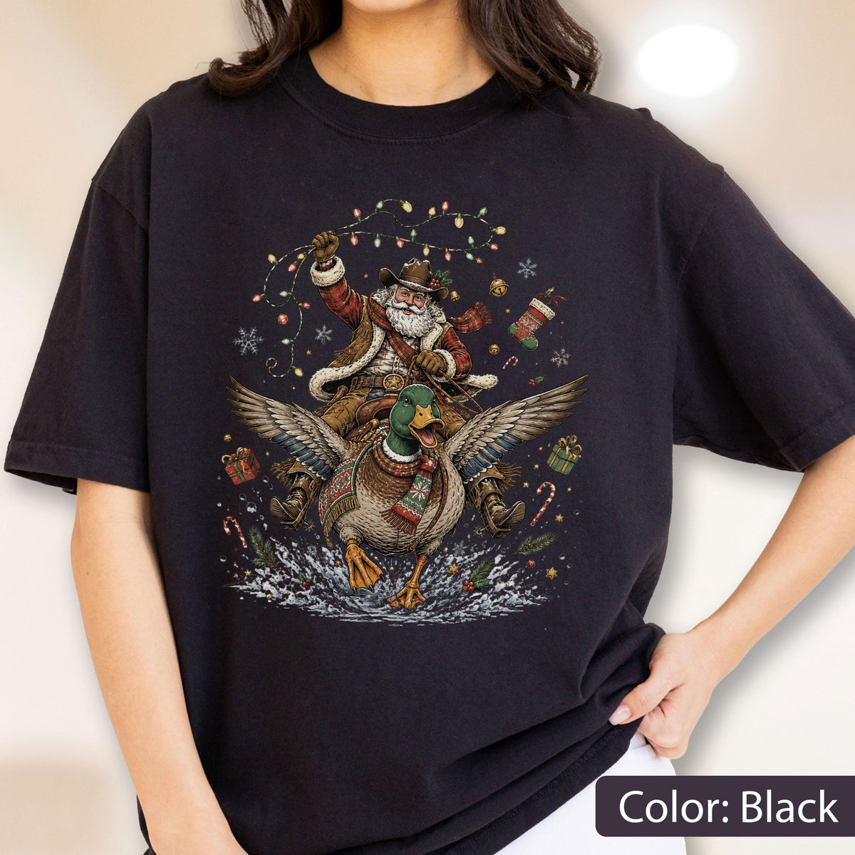 May include: A black t-shirt featuring a graphic print of Santa Claus wearing a cowboy hat and riding a mallard duck. The design includes colorful Christmas lights, snowflakes, wrapped gifts, a stocking, and candy canes surrounding the figures.