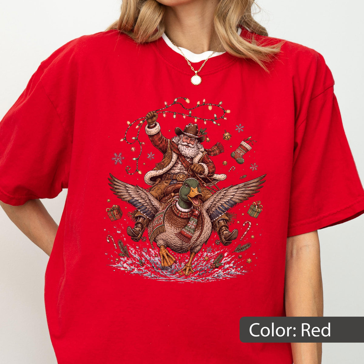 May include: A bright red short-sleeved t-shirt featuring a graphic print of Santa Claus wearing a cowboy hat and riding a flying mallard duck. The illustration includes festive elements like string lights, snowflakes, gift boxes, and a stocking.