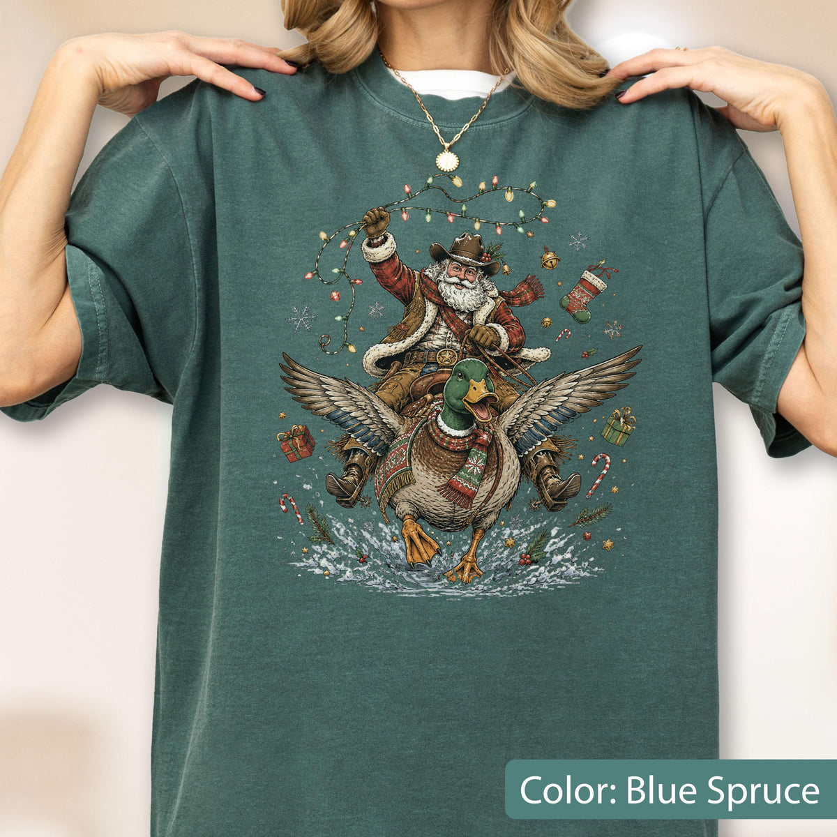May include: A blue spruce t-shirt featuring a graphic of Santa Claus wearing a cowboy hat and riding a flying mallard duck. The design includes a string of colorful Christmas lights, a stocking, gift boxes, and candy canes against a snowy backdrop.