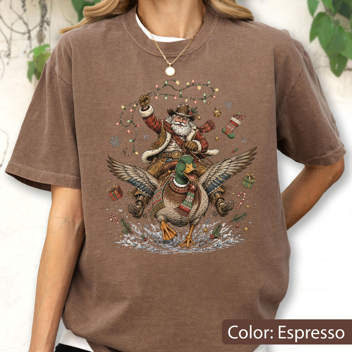 May include: An espresso brown t-shirt featuring a graphic print of Santa Claus wearing a cowboy hat and riding a mallard duck. The design includes Christmas lights, snowflakes, wrapped gifts, a stocking, and a candy cane surrounding the figures.