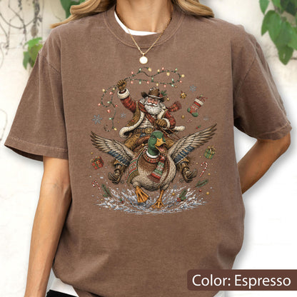 May include: An espresso brown t-shirt featuring a graphic print of Santa Claus wearing a cowboy hat and riding a mallard duck. The design includes Christmas lights, snowflakes, wrapped gifts, a stocking, and a candy cane surrounding the figures.