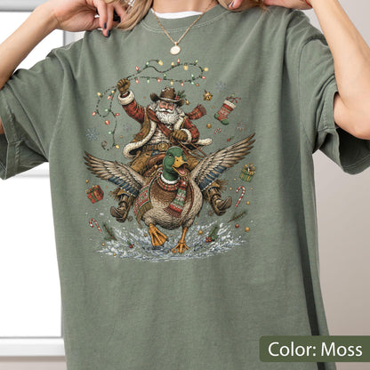 May include: A moss green t-shirt featuring a graphic illustration of Santa Claus wearing a cowboy hat and scarf, riding a flying mallard duck. The scene includes Christmas lights, a stocking, candy canes, and gift boxes on a snowy background.