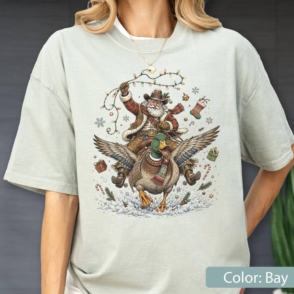 May include: A light sage green t-shirt featuring a graphic print of Santa Claus wearing a cowboy hat and vest while riding a flying mallard duck. The design includes Christmas lights, snowflakes, wrapped gifts, and candy canes in the scene.