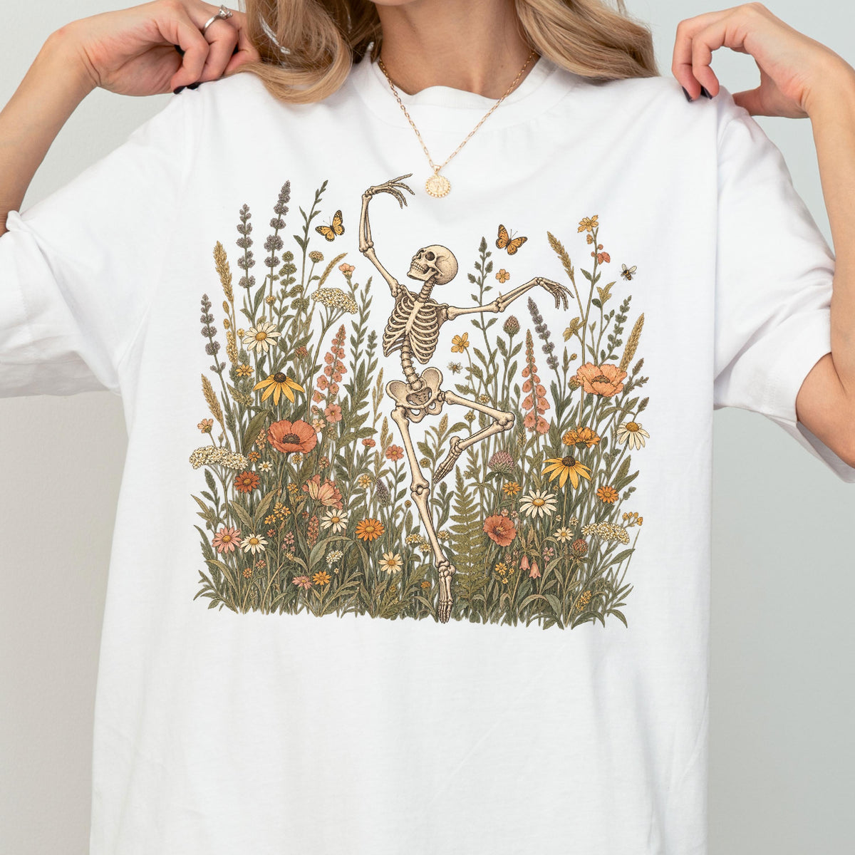 May include: A white t-shirt featuring a graphic print of a skeleton dancing amidst a field of colorful wildflowers, including poppies, daisies, and lavender, with small butterflies and bees scattered throughout the floral scene.