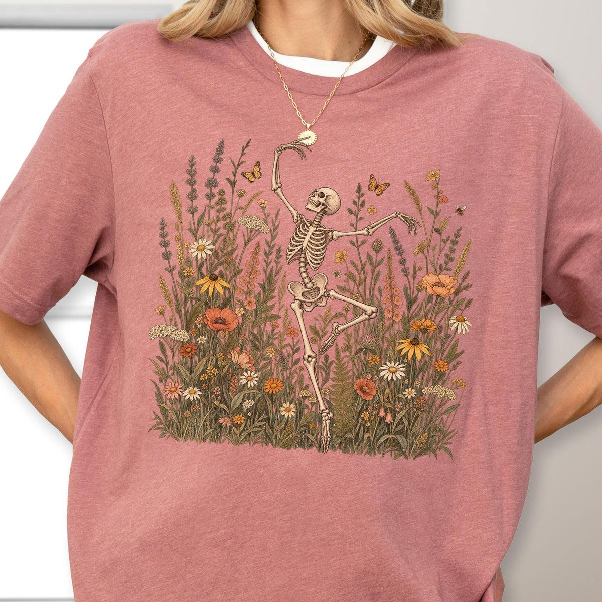 May include: A mauve short-sleeved t-shirt featuring a graphic print of a skeleton dancing in a field of wildflowers and butterflies. The skeleton is positioned centrally, surrounded by various orange, yellow, and white flowers and green foliage.