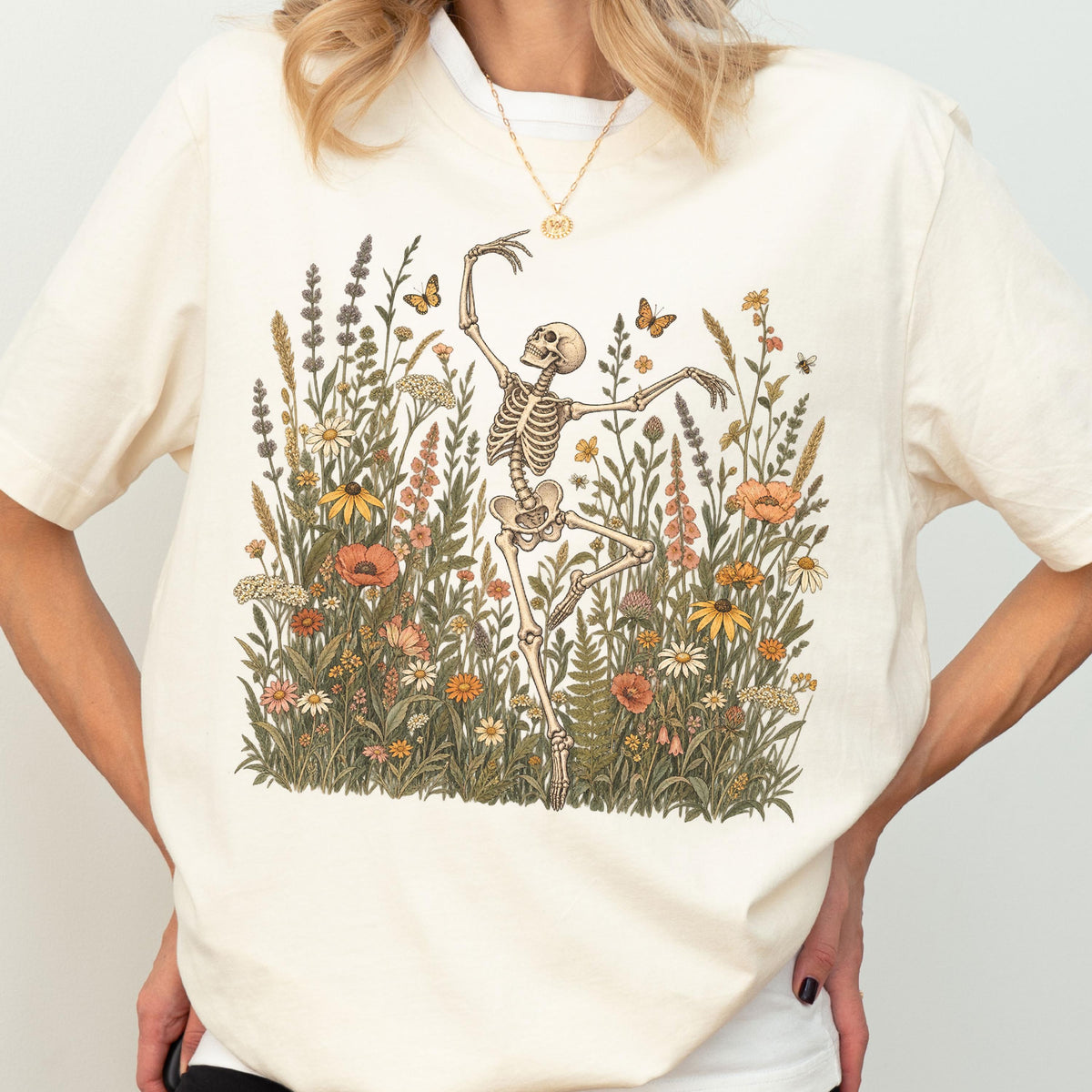 May include: A cream-colored short-sleeve t-shirt featuring a graphic print of a skeleton dancing amidst a field of colorful wildflowers, tall grasses, and small butterflies. The design is centered on the chest area of the garment.