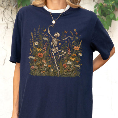 May include: A navy blue short-sleeve t-shirt featuring a graphic print of a skeleton dancing amidst a field of colorful wildflowers, including daisies, poppies, and various green foliage, with small butterflies scattered throughout the scene.