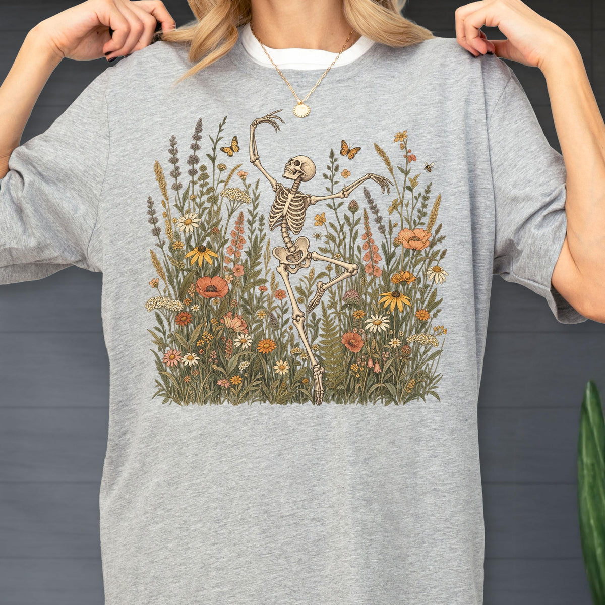 May include: A heather gray t-shirt featuring a graphic print of a skeleton dancing amidst a field of colorful wildflowers and butterflies. The design includes various botanical elements in shades of orange, yellow, and green, centered on the chest.