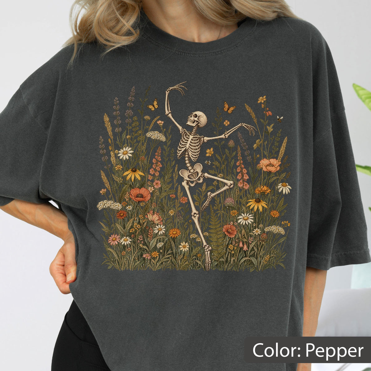 May include: A charcoal gray t-shirt featuring a graphic print of a human skeleton dancing amidst a field of colorful wildflowers and small butterflies. The skeleton is positioned in the center, surrounded by various botanical elements in shades of orange, yellow, and green.