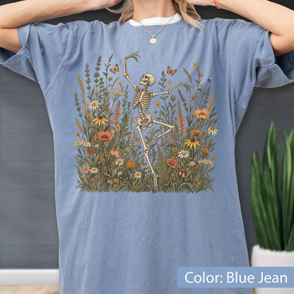May include: A blue jean colored t-shirt featuring a graphic print of a skeleton dancing in a field of wildflowers. The illustration includes various colorful flowers, green foliage, and small butterflies surrounding the central skeleton figure.