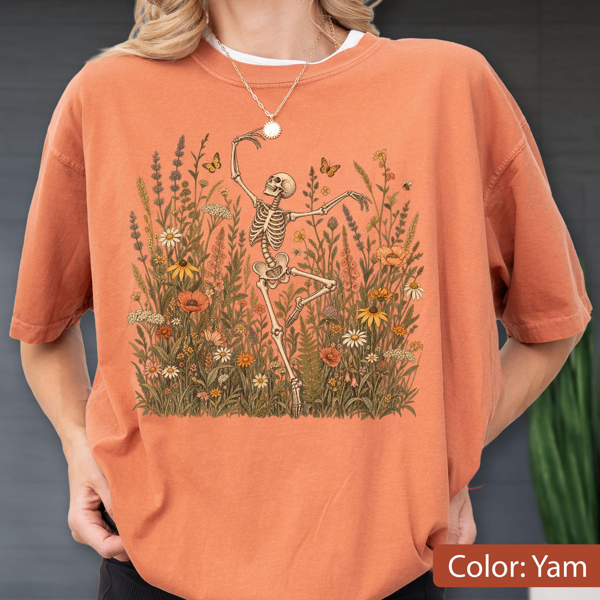 May include: A yam-colored t-shirt featuring a graphic print of a skeleton dancing amidst a field of wildflowers and butterflies. The design includes various botanical elements in shades of green, yellow, orange, and white, centered on the chest.