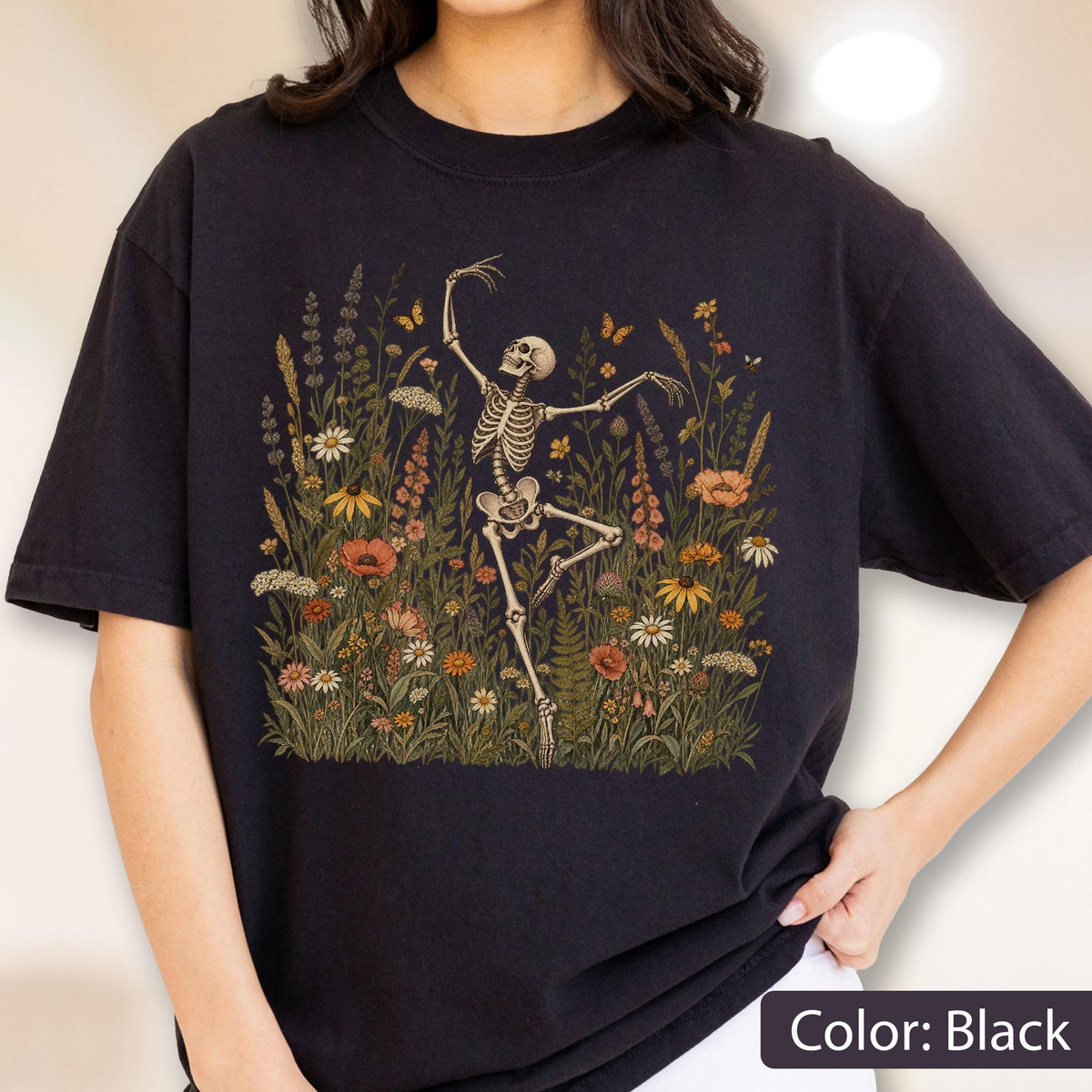 May include: A black t-shirt featuring a graphic print of a skeleton dancing amidst a field of colorful wildflowers and small butterflies. The illustration includes various botanical elements in shades of orange, yellow, white, and green.