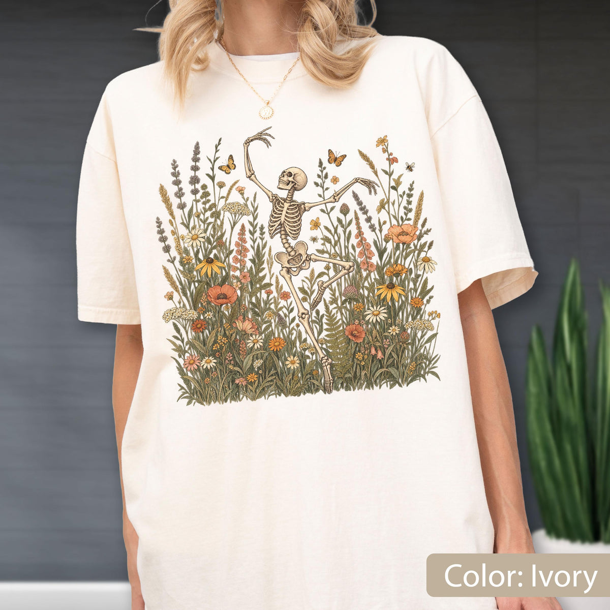 May include: An ivory short-sleeve t-shirt featuring a graphic print of a skeleton dancing amidst a field of wildflowers, including poppies, daisies, and lavender, with small butterflies and bees scattered throughout the floral arrangement.