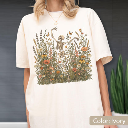 May include: An ivory short-sleeve t-shirt featuring a graphic print of a skeleton dancing amidst a field of wildflowers, including poppies, daisies, and lavender, with small butterflies and bees scattered throughout the floral arrangement.