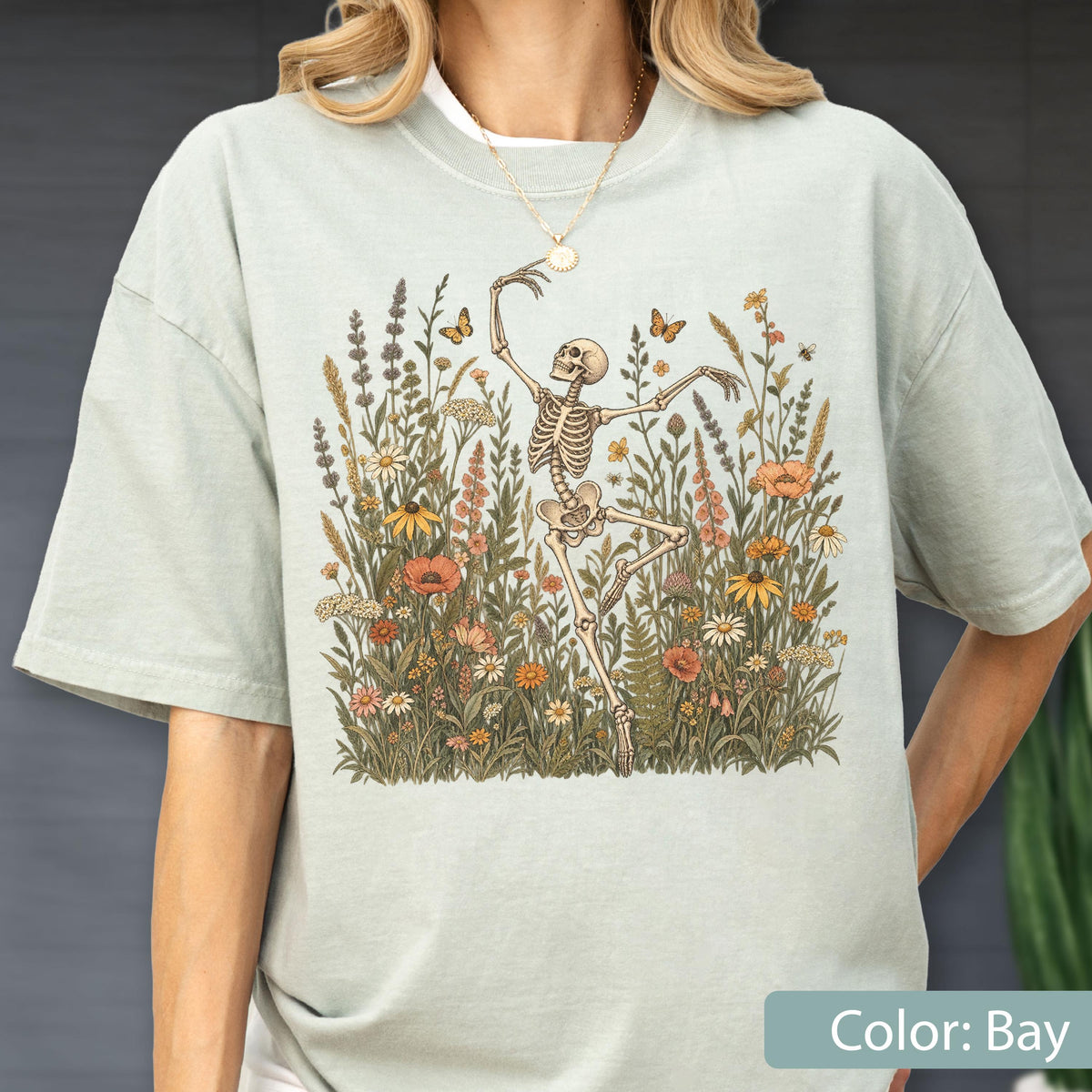 May include: A light sage green short-sleeve t-shirt featuring a central graphic of a skeleton dancing amidst a field of wildflowers and butterflies. The illustration includes various botanical elements in muted earth tones, centered on the chest.