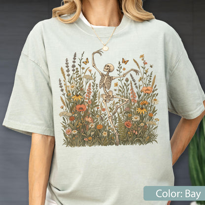 May include: A light sage green short-sleeve t-shirt featuring a central graphic of a skeleton dancing amidst a field of wildflowers and butterflies. The illustration includes various botanical elements in muted earth tones, centered on the chest.