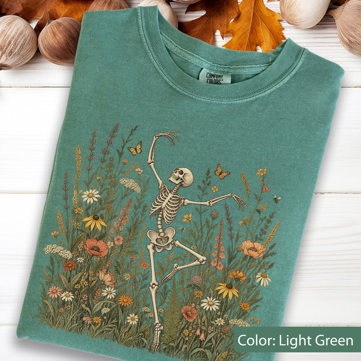 May include: A light green t-shirt featuring a graphic print of a skeleton dancing amidst a field of colorful wildflowers and butterflies. The shirt has a crew neckline and a Comfort Colors brand label printed on the inside of the collar area.