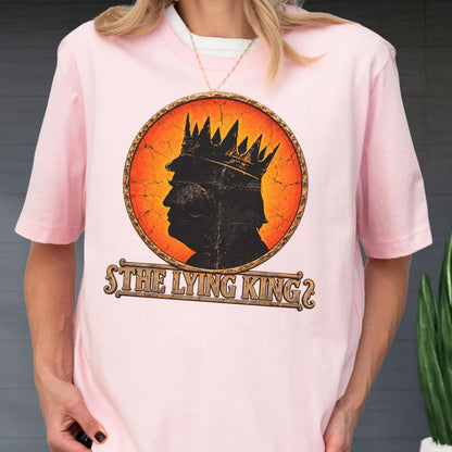 May include: A light pink short-sleeved t-shirt featuring a circular graphic with a black silhouette of a person wearing a crown against an orange and yellow cracked background. Below the circle, the text reads THE LYING KING in a stylized font.