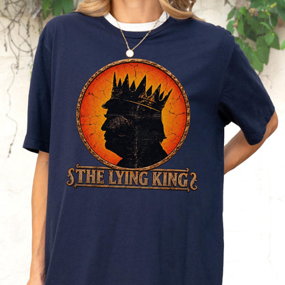 May include: A navy blue t-shirt featuring a circular graphic with a distressed orange and red background. The graphic shows a black silhouette of a person wearing a crown. Below the circle, the text reads THE LYING KING in a stylized font.