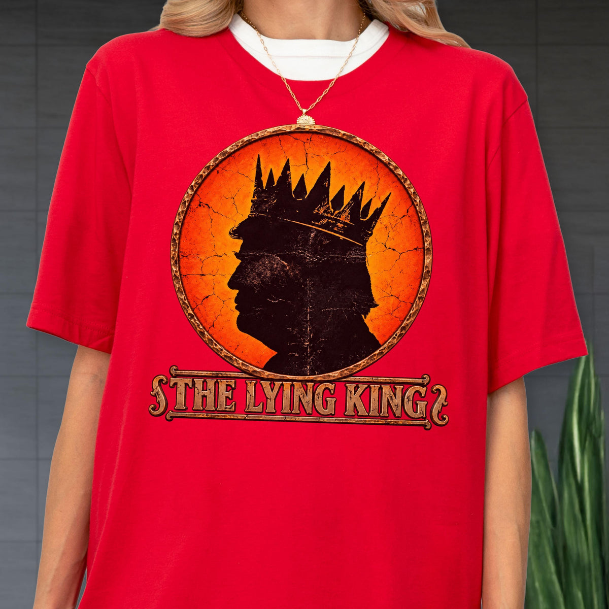 May include: A bright red short-sleeved t-shirt featuring a circular graphic with a cracked orange background. Inside the circle is a black silhouette of a person wearing a crown. Below the circle, the text reads THE LYING KING in a bold font.