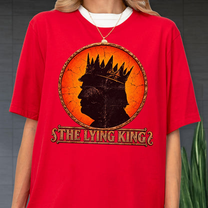 May include: A bright red short-sleeved t-shirt featuring a circular graphic with a cracked orange background. Inside the circle is a black silhouette of a person wearing a crown. Below the circle, the text reads THE LYING KING in a bold font.