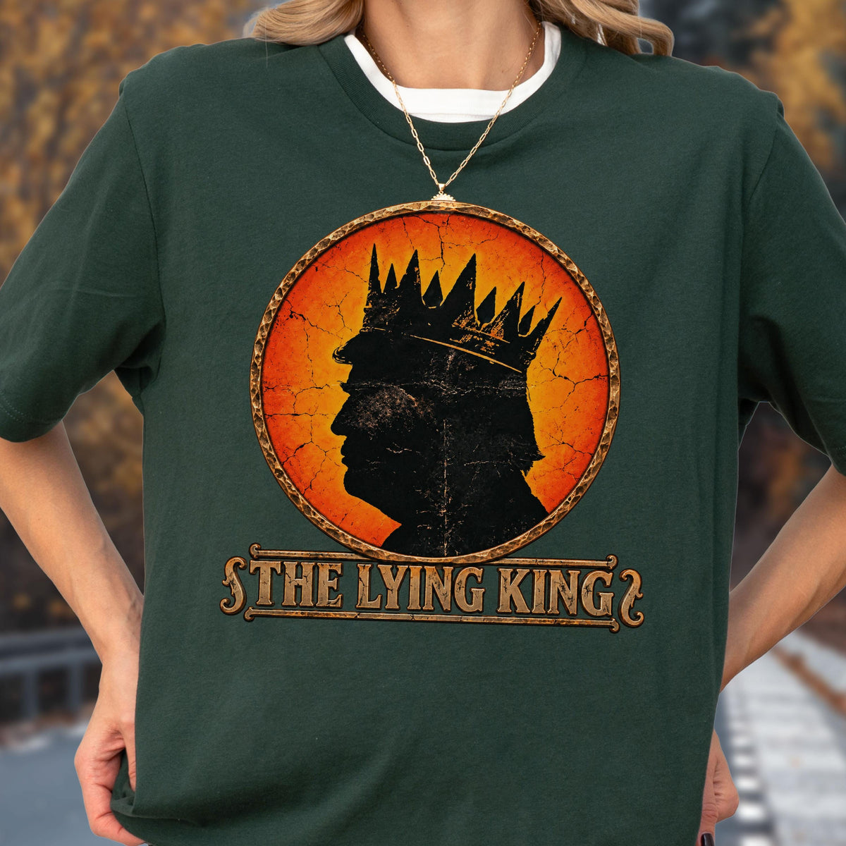 May include: A dark green short-sleeve t-shirt featuring a circular graphic with a distressed orange and yellow background. The graphic shows a black silhouette of a person wearing a crown, with the text THE LYING KING printed below in gold.