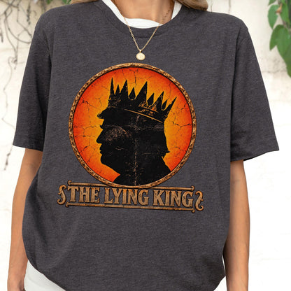 May include: A charcoal gray short-sleeve t-shirt featuring a circular graphic with a distressed orange and yellow background. The graphic displays a black silhouette of a person wearing a crown, with the text THE LYING KING printed below.